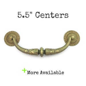 May include: Antique brass-colored drawer pull with a decorative floral design. The pull has a 5.5-inch center-to-center measurement and includes the text "5.5" Centers" and "More Available".