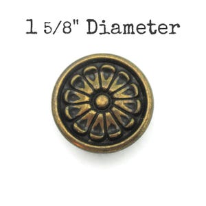 May include: A round, antique brass-colored knob with a floral design. The knob has a diameter of 1 5/8 inches. The design features a central circle surrounded by petal-like shapes. The knob is likely for furniture or decorative use.