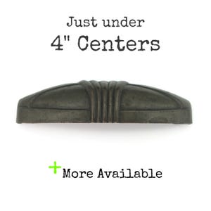 May include: A dark metallic drawer pull with a curved, ribbed centre design. The text above reads Just under 4" Centers and the text below reads + More Available. The hardware features a smooth, matte finish and a symmetrical, elongated shape.