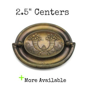 May include: An oval brass drawer pull with a drop handle, featuring a central embossed design of two cornucopias filled with fruit and foliage. The hardware is labeled with the text 2.5" Centers and More Available.