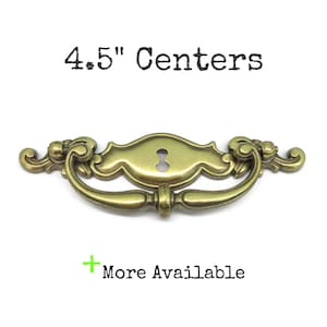 May include: A brass-toned metal drawer pull with a central keyhole escutcheon and a swinging handle. The text 4.5" Centers is printed above the hardware, and the text + More Available is printed below the hardware on a white background.
