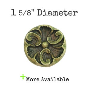 May include: A decorative, antique-style button with a floral design. The button is 1 5/8" in diameter and has a bronze finish with green accents. The text "More Available" is below the button.