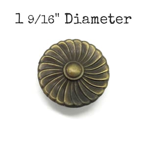May include: A round, antique brass-colored cabinet knob with a floral design. The knob has a diameter of 1 9/16 inches. The center of the knob is a small, raised circle, and the outer edge has a series of curved lines.