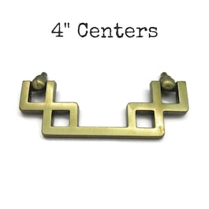 May include: A brass-toned cabinet pull with a geometric design. The pull has a rectangular shape with square cutouts and a decorative knob on each end. The text "4" Centers" is above the pull.