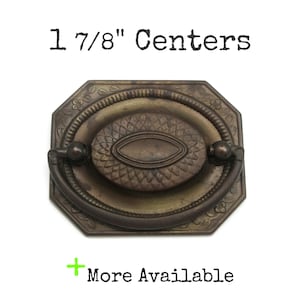 May include: An antique brass drawer pull with an octagonal backplate featuring a floral border and a central oval medallion. The hardware includes a swinging bail handle and is labeled with the text 1 7/8" Centers and + More Available.