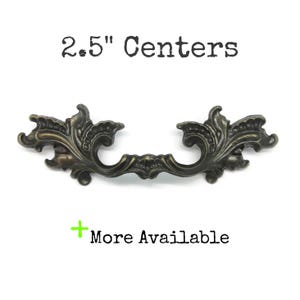 May include: A dark bronze-toned metal drawer pull with an ornate, scrolling design. The text 2.5" Centers is printed above the handle, and the text + More Available is printed below the handle on a plain white background.