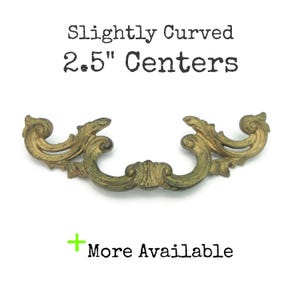 May include: An antique-style brass drawer pull with a slightly curved design. The pull has a 6.35 cm centre-to-centre measurement and features decorative scrollwork. The text "Slightly Curved 2.5" Centers" is above the pull, and "More Available" is below.