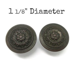 May include: Two round, dark bronze-toned metal knobs featuring a central floral relief pattern surrounded by a beaded border. The text 1 1/8" Diameter is printed above the knobs, indicating their size. These circular hardware pieces have a matte finish.