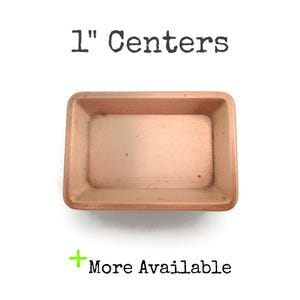 May include: A rectangular copper-colored drawer pull with a recessed center. The text "1" Centers" is above the pull, and "More Available" is below. The pull measures 1 inch in length.