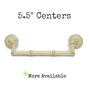 May include: Off-white cabinet pull with a bamboo-style handle and decorative circular end pieces. The text "5.5" Centers" is above the pull, and "More Available" is below. The hardware is designed for furniture.