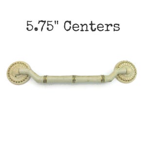 May include: Off-white cabinet pull with a bamboo-style handle and decorative circular ends. The pull has brass-coloured accents and is labelled "5.75" Centers". This hardware is suitable for drawers and cabinets.