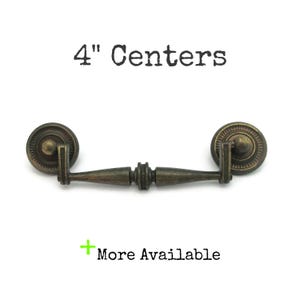 May include: Antique bronze drawer pull with a 4-inch center-to-center measurement. The hardware features a decorative design with circular end pieces and a central handle. The text "4" Centers" and "More Available" are visible.