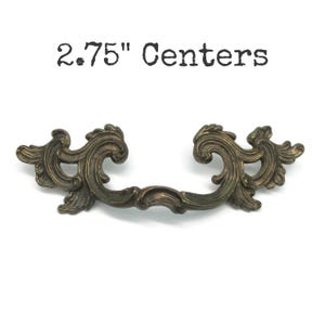May include: A bronze-toned metal drawer pull with an ornate, scrolling leaf design and a curved handle. The text 2.75" Centers is printed above the hardware, indicating the distance between the mounting points for this furniture handle.