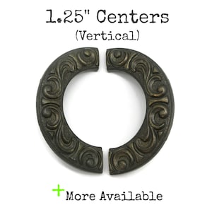 May include: Two dark bronze-toned metal drawer pulls with an ornate, curved scrollwork pattern. The text above reads 1.25" Centers (Vertical) and the text below reads + More Available. The hardware features a weathered, textured finish.