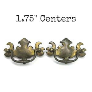 May include: Two antique brass drawer pulls with a decorative design. The pulls have a curved handle and ornate detailing. The text "1.75" Centers" is printed above the pulls.