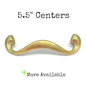 May include: A brass-coloured metal drawer pull with a curved, wave-like shape and scrolled ends. The text 5.5" Centers is printed above the handle, and the text + More Available is printed below the handle in black lettering.