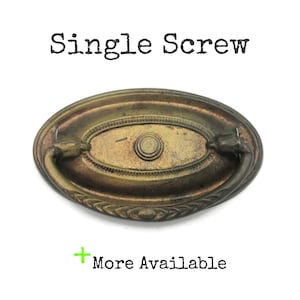 May include: Antique brass oval drawer pull with a central circular design and curved handle. The text "Single Screw" is above the pull, and "More Available" is below. The pull has a decorative border.