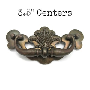 May include: Antique brass drawer pull with a decorative floral design. The pull measures 3.5 inches (8.89 cm) between the centers of the mounting holes. The handle has a curved design and is mounted on a decorative backplate.