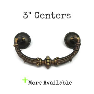 May include: A dark bronze-toned metal drawer pull with a curved, ornate handle and two circular mounting plates. The text 3" Centers is printed above the handle, and the text + More Available is printed below the handle on a white background.