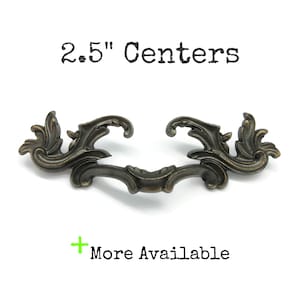 May include: A dark bronze-toned metal drawer pull with ornate, scrolling leaf-like details and a curved handle. The text 6.35 cm Centers is printed above the handle, and a green plus sign with the text More Available is printed below.