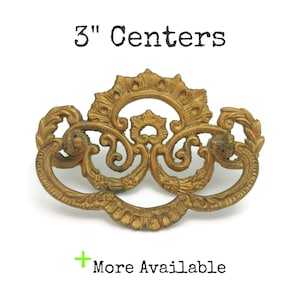 May include: Ornate gold-colored drawer pull with a decorative design. The pull has a 3-inch center-to-center measurement and features scrolling details and a floral motif. The text "3" Centers" is above the pull, and "More Available" is below.