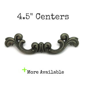 May include: An antique bronze-colored drawer pull with an ornate, scrolling design. The text "4.5" Centers" is above the pull, and "More Available" is below. The hardware measures 4.5 inches across.