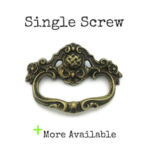 May include: An antique brass-toned drawer pull with an ornate design. The pull features a central woven pattern and curved handle. The text "Single Screw" is above the pull, and "More Available" is below it.