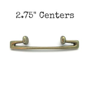 May include: A brass-toned metal drawer pull with a horizontal bar and two rounded mounting points, featuring the text 2.75" Centers above the hardware. The handle has a smooth, slightly weathered finish with a distinct geometric shape.