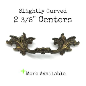 May include: A bronze-toned metal drawer pull with an ornate, leaf-like design and a slightly curved shape. The text above the pull reads Slightly Curved 2 3/8" Centers, and the text below reads + More Available.