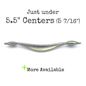 May include: A silver-toned, curved cabinet pull with a 5.5 inch center-to-center measurement. The text "Just under 5.5" Centers (5 7/16")" is visible above the pull.