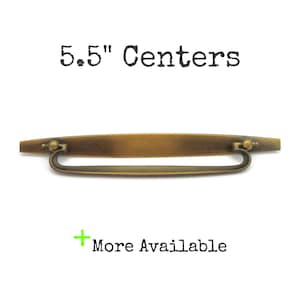 May include: Antique brass-toned drawer pull with a curved handle. The text "5.5" Centers" is above the pull, and "More Available" is below. The pull measures 5.5 inches in length.