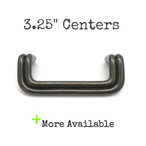 May include: A dark metallic cabinet pull with a double-bar design and a smooth finish. The text 3.25" Centers is printed above the handle, and the text + More Available is printed below the handle in black lettering on a white background.