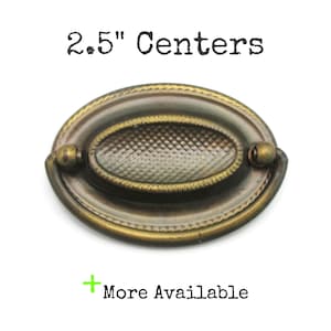 May include: Oval antique brass drawer pull with a textured centre and decorative trim. The text "2.5" Centers" is above the pull. The text "More Available" is below the pull.