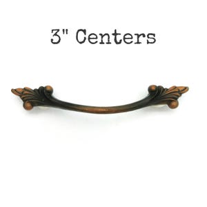 May include: A bronze-colored cabinet pull with a curved design and decorative leaf accents. The pull has a 3-inch center-to-center measurement. The text "3" Centers" is above the pull.