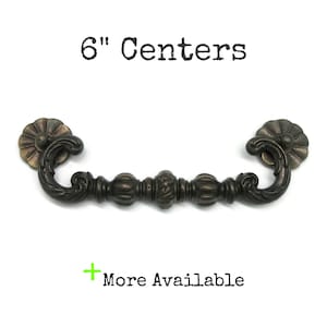 May include: A dark bronze-toned metal drawer pull with a curved, ornate design and circular mounting plates. The text above the handle reads 6" Centers, and the text below reads + More Available, indicating the hardware's specific mounting measurement.