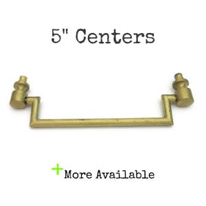 May include: A brass-toned drawer pull with a 5-inch center-to-center measurement. The pull has a rectangular bar with decorative end pieces. The text "5" Centers" and "More Available" are visible.