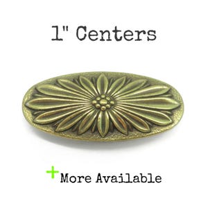 May include: An oval-shaped brass-toned metal drawer pull featuring a raised floral design with a central cluster of small dots surrounded by elongated petals. The text 1" Centers is printed above the pull, and + More Available is printed below.