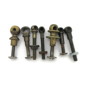 May include: Seven antique metal ball joint screws are displayed, each with a threaded shaft and a ball-shaped head with a hole. The screws have varying sizes, colours, and designs, including brass and steel finishes. They are set against a white backdrop.