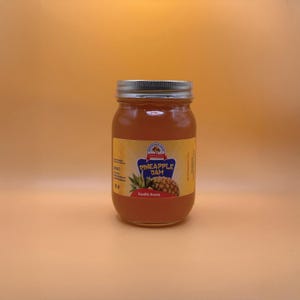 May include: A jar of pineapple jam with a silver lid and a yellow label. The label features a pineapple illustration and the words "Pineapple Jam". The jam has a golden-orange color.