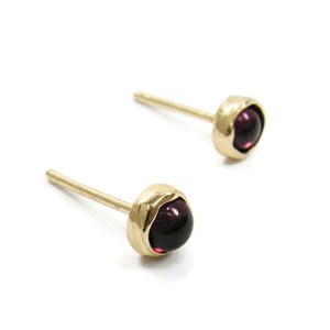 May include: A pair of gold stud earrings with round, deep purple gemstones. The earrings have a simple, elegant design, with the gemstones set in a bezel setting. The posts are thin and gold. The earrings are displayed on a white background.