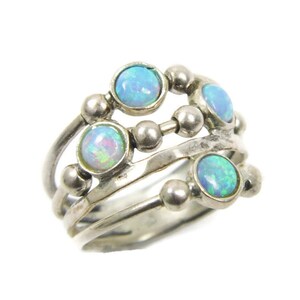 May include: A silver ring featuring multiple bands and four round, iridescent opal stones. The ring has a unique design with small silver beads connecting the bands. The opals display a range of blue and green hues.