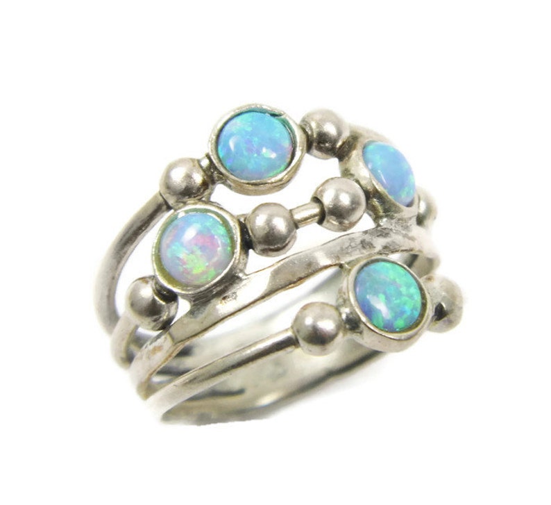 May include: A silver ring featuring multiple bands and four round, iridescent opal stones. The ring has a unique design with small silver beads connecting the bands. The opals display a range of blue and green hues.
