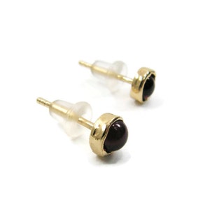 May include: A pair of gold stud earrings with dark red, round gemstones. The earrings have a bezel setting and are shown with clear earring backs. The studs are on a white background.