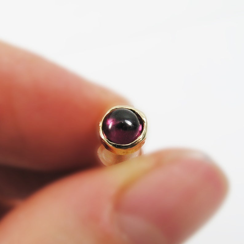 May include: Close-up of a small, gold-colored stud earring with a round, deep red gemstone. The gemstone is set in a gold bezel, creating a simple yet elegant design. The earring is held between fingers against a white background.
