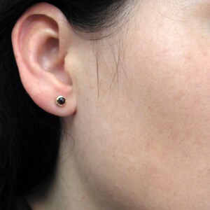 May include: A close-up of a person's ear with a small, round stud earring. The earring features a dark, possibly black, gemstone set in a gold-colored metal. The earring is a simple, classic design.