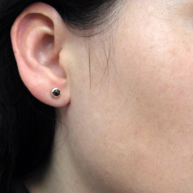 May include: A close-up of a person's ear with a small, round stud earring. The earring features a dark, possibly black, gemstone set in a gold-colored metal. The earring is a simple, classic design.