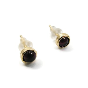 May include: A pair of stud earrings with a gold-colored bezel setting. Each earring features a round, dark red gemstone. The earrings have a post and a clear, plastic backing for secure wear.
