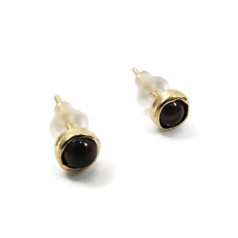 May include: A pair of stud earrings with a gold-colored bezel setting. Each earring features a round, dark red gemstone. The earrings have a post and a clear, plastic backing for secure wear.