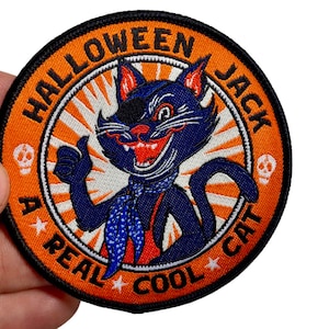 Op de afbeelding: Een geborduurde Halloween-patch met een oranje achtergrond en een zwarte rand. De patch toont een zwarte kat met een blauw lichaam en een rode bandana. De tekst "A Real Cool Cat" is in een cirkel rond de kat geschreven. De tekst "Halloween Jack" is in een cirkel boven de kat geschreven.