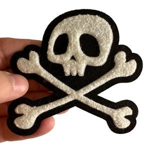May include: A black skull and crossbones patch with a white, textured skull and crossbones design. The patch is held in a hand and is likely an iron-on or sew-on patch for clothing or accessories.